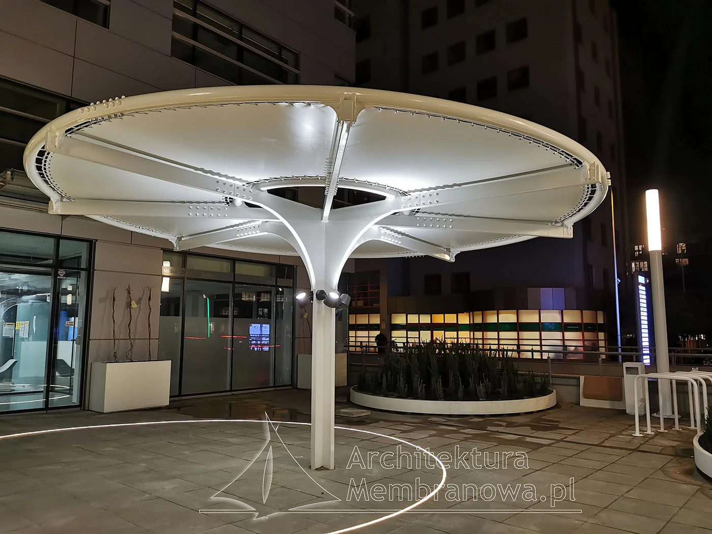 Podświetlone okrągłe zadaszenie membranowe na centralnym maszcie na dziedzińcu biurowca, nocą